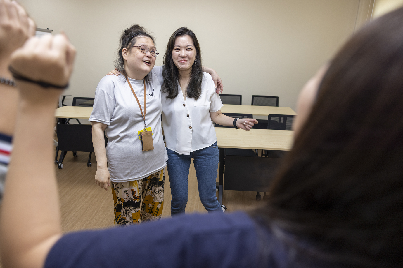 Two women in casual attire pose with arms around each other in a classroom setting.